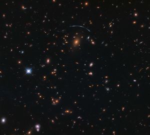 Black background dotted with galaxies. A reddish galaxy at image center with a bluish-white arc above it.
